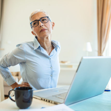 Portrait of senior stressed woman sitting at home office desk in front of laptop, touching aching back with pained expression, suffering from back pain after working on laptop