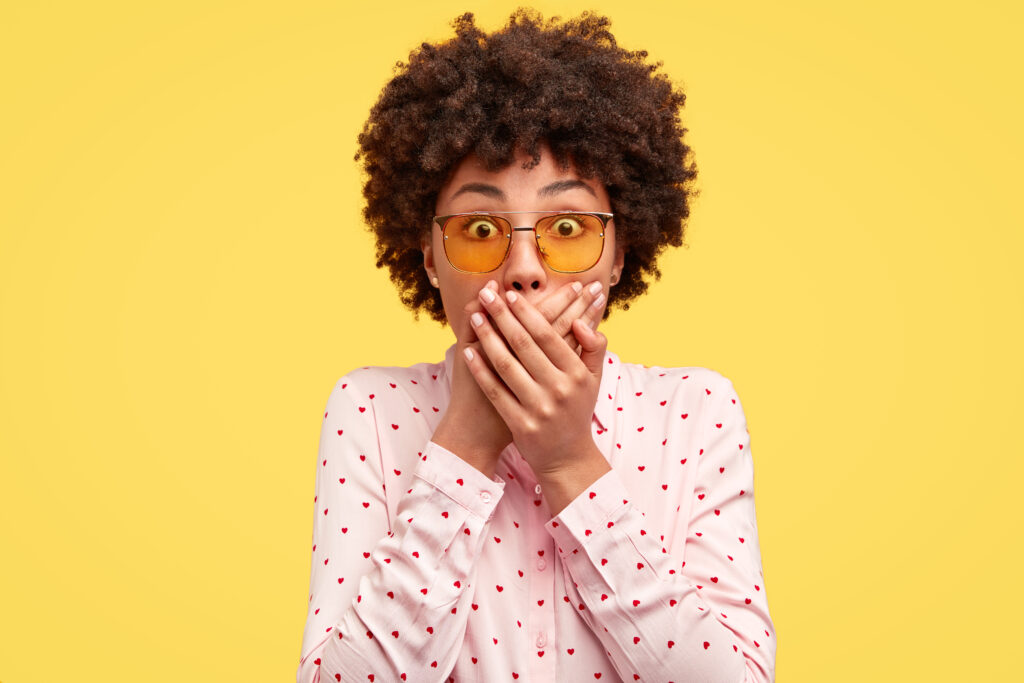 Stunned dark skinned woman with curly hair, covers both hands on mouth, afraids of something astonishing, dressed in fashionable shirt, isolated over yellow background, stands speechless indoor