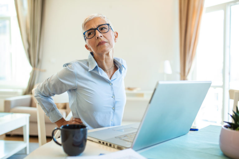 Portrait of senior stressed woman sitting at home office desk in front of laptop, touching aching back with pained expression, suffering from back pain after working on laptop