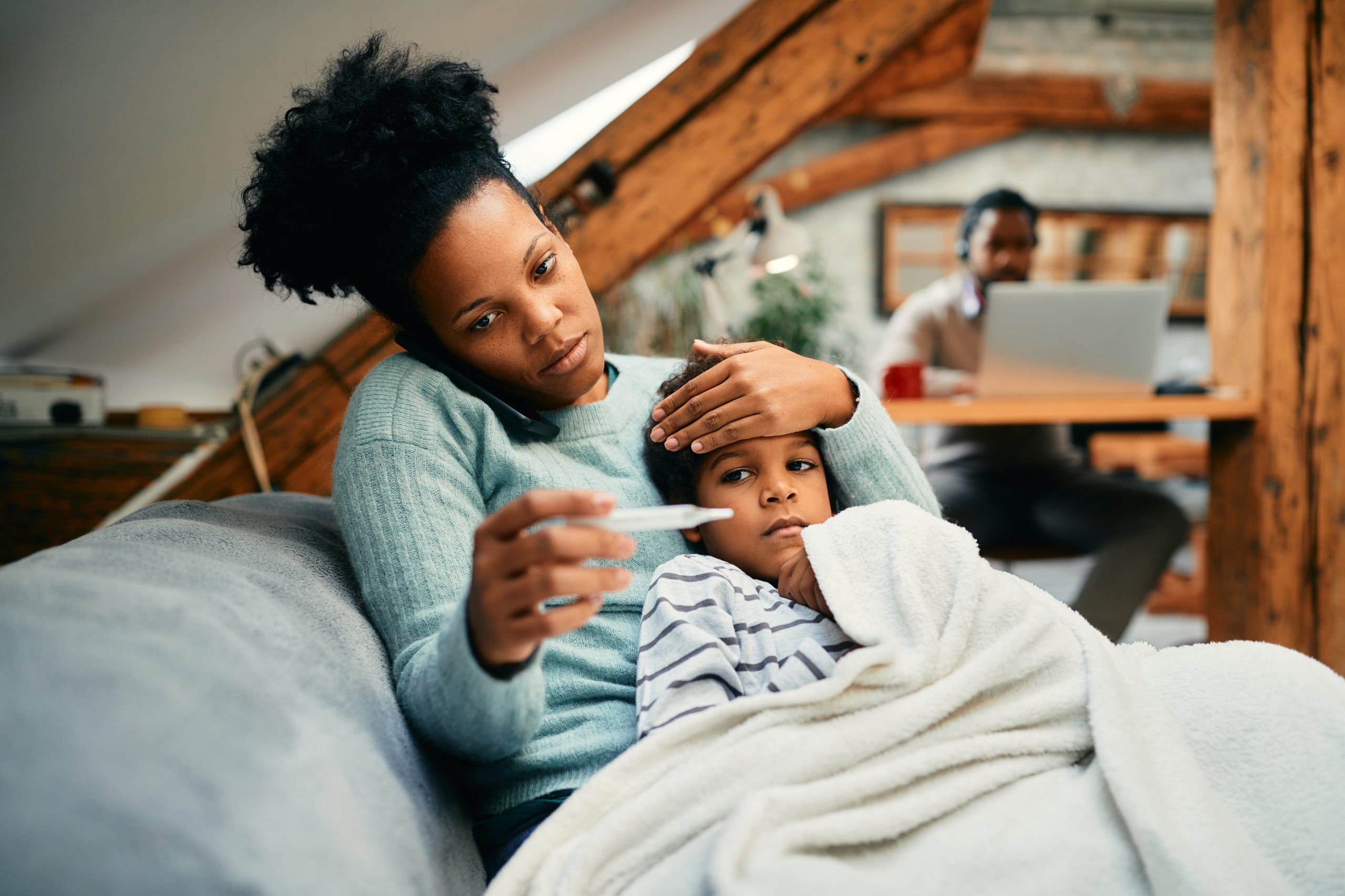 Worried black mother talks on the phone while measuring son's temperature at home.