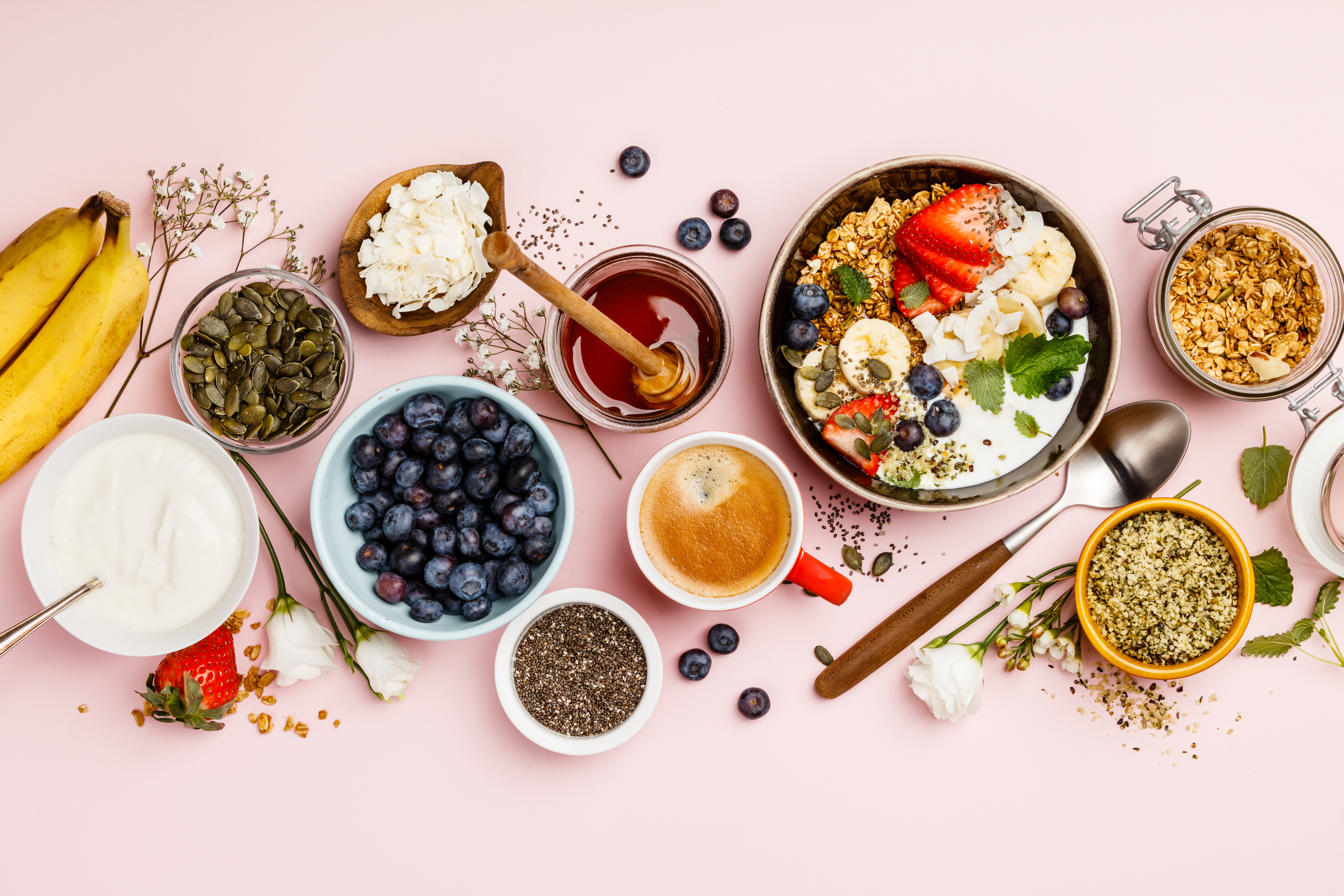 Healthy breakfast set. Oat granola with fresh berries, banana, yogurt, maple syrup, seeds and mint leaves with cup of coffee on pink background, flat lay, top view